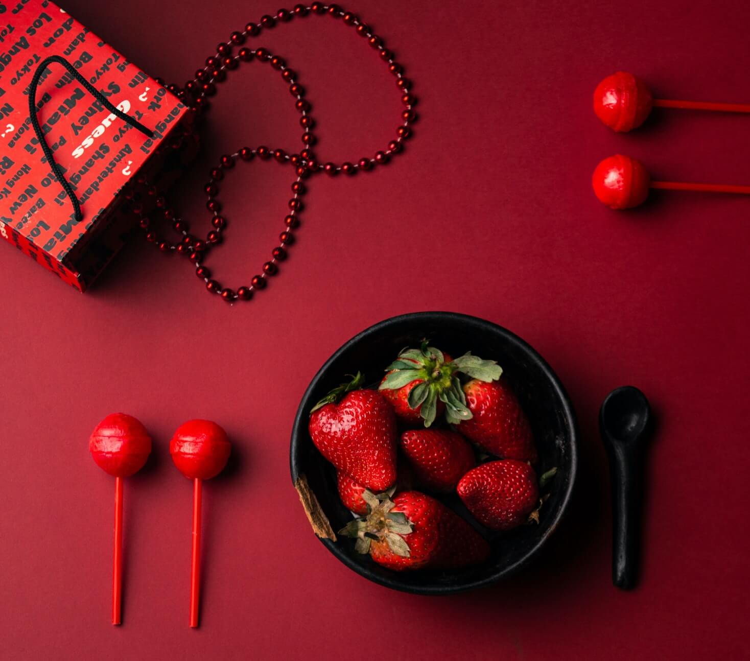 collection d'objets rouges sur fond rouge : sac en papier rouge, collier de perles rouges, sucette et bonbons rouges, fraises rouges dans un bol noir
